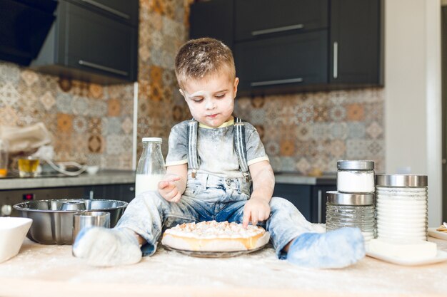 Drôle enfant assis sur la table de la cuisine dans une cuisine roustique jouant avec de la farine et dégustant un gâteau.