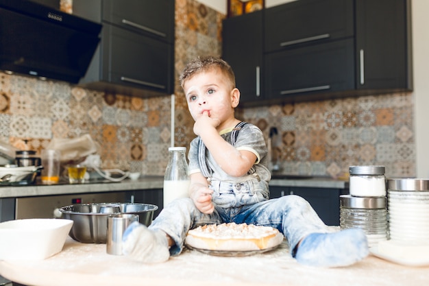 Drôle enfant assis sur la table de la cuisine dans une cuisine roustique jouant avec de la farine et dégustant un gâteau.