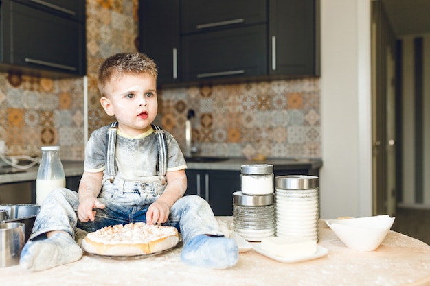 Drôle enfant assis sur la table de la cuisine dans une cuisine roustique jouant avec de la farine et dégustant un gâteau.