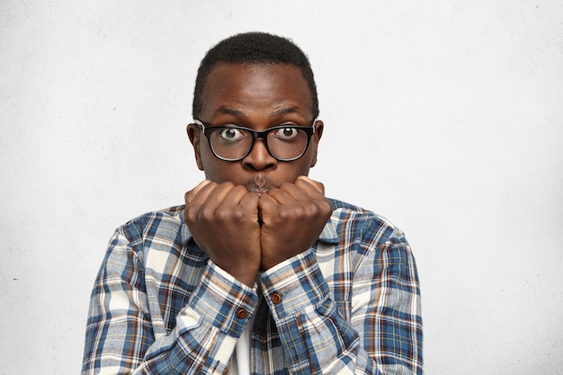 Drôle d'étudiant afro-américain aux yeux pop en lunettes se sentant nerveux et effrayé avant les examens à l'université, gardant les mains dans les poings sur son visage. Homme noir à la peur et effrayé par quelque chose