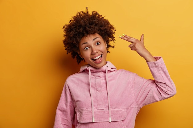 Drôle de femme afro-américaine tient la tempe du pistolet à doigt, incline la tête, fait un geste de suicide, sourit largement, se trompe, porte un sweat-shirt, pose contre le mur jaune. Concept de langage corporel