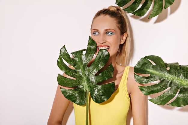 Drôle de femme aux cheveux blonds avec un maquillage lumineux en maillot de bain motif banane jaune mordant de grandes feuilles vertes sur fond blanc