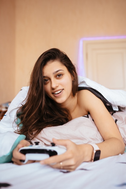 Drôle femme couchée dans son lit et jouer au jeu vidéo, tenant le contrôleur