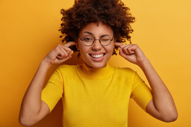 Drôle de femme à la peau sombre se branche les doigts dans les oreilles, ne peut pas se concentrer à cause du bruit dans un endroit bondé, fait un clin d'œil et montre des dents blanches, porte des lunettes et un t-shirt, isolé sur un mur jaune