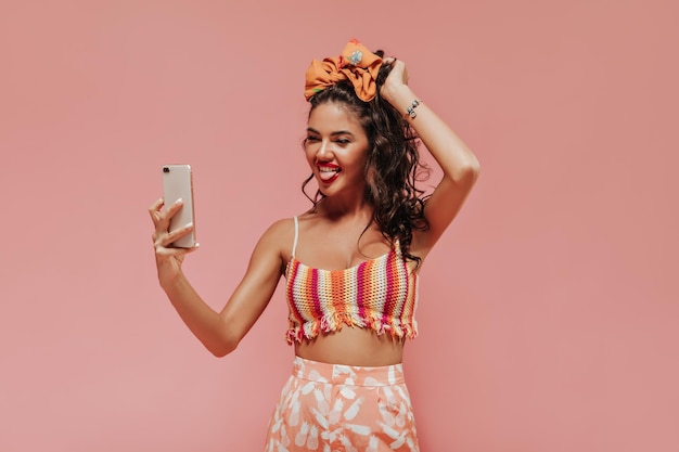 Une drôle de fille bouclée avec un bandeau élégant et des accessoires dans un haut coloré et un pantalon à imprimé ananas fait une photo sur fond rose