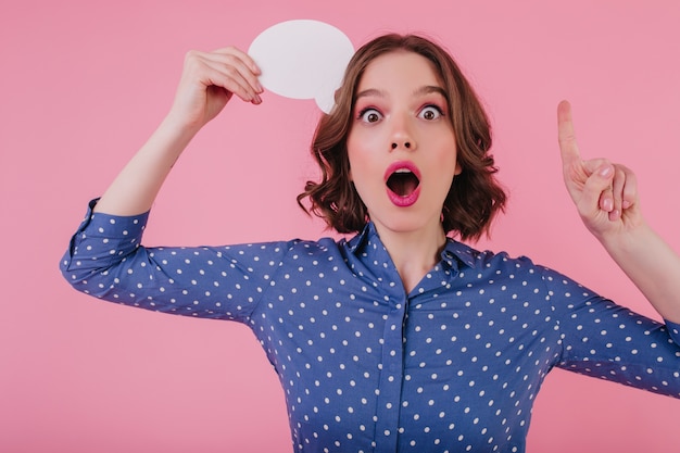 Drôle de fille étonnée a une idée. Modèle féminin aux cheveux courts enthousiaste pensant à quelque chose sur un mur rose.