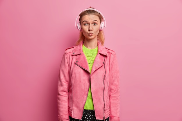 Une Drôle De Fille Européenne Garde Les Lèvres Rondes, écoute De La Musique Dans Des écouteurs, Choisit Une Chanson Dans Une Liste De Lecture, Est En Route Pour L'école, Vêtue D'une Veste Rose à La Mode, Aime La Chanteuse Préférée