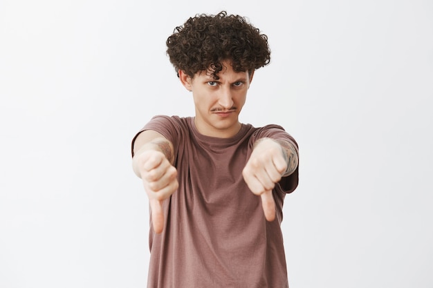 Photo gratuite drôle de gars juif mécontent et peu impressionné avec des cheveux bouclés et une moustache regardant sous le front avec un regard fou montrant les pouces vers le bas dans un geste de dégoût donnant des commentaires négatifs ou une réponse