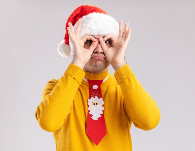 Drôle et heureux jeune homme en col roulé jaune et bonnet de noel avec cravate drôle regardant à travers les doigts faisant le geste binoculaire debout sur fond blanc