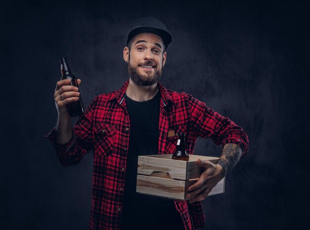 Un drôle de hipster ivre barbu tient une boîte en bois avec des bouteilles de bière, sur un fond sombre.