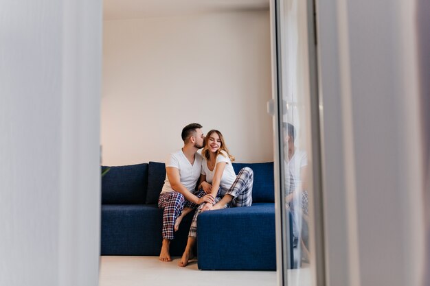 Drôle d'homme aux pieds nus en pyjama embrassant sa femme. Portrait intérieur de couple marié paresseux appréciant le matin.