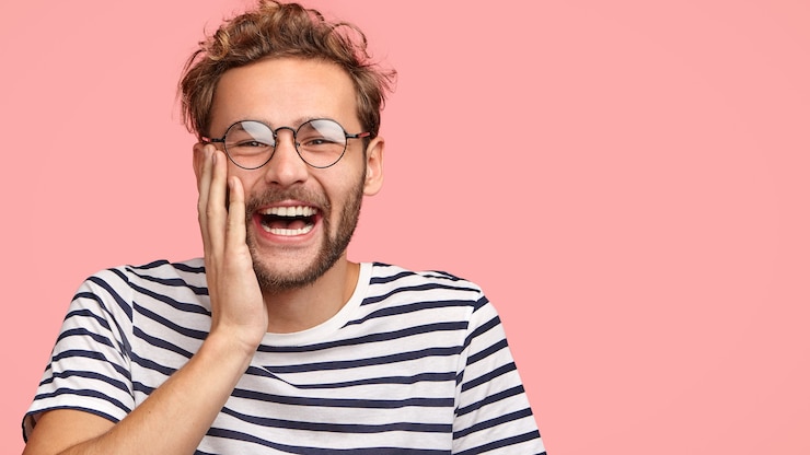 Photo gratuit un drôle d'homme bouclé rit joyeusement, touche les joues, regarde un programme intéressant, vêtu d'un t-shirt à rayures décontracté, se tient contre le mur rose.