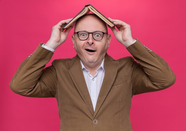 Drôle homme chauve d'âge moyen en costume portant des lunettes tenant un livre ouvert sur sa tête souriant à la surprise debout sur le mur rose