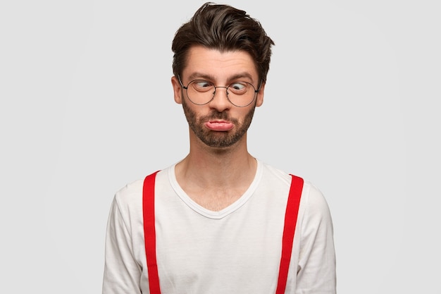 Photo gratuite un drôle d'homme mal rasé croise les yeux et serre les lèvres, fait la grimace, a une expression comique, fou à l'intérieur contre un mur blanc. un homme caucasien barbu élégant n'a rien à faire.