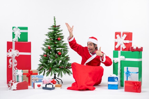 Drôle jeune adulte habillé en père Noël avec des cadeaux et arbre de Noël décoré assis sur le sol pointant au-dessus de faire un geste parfait sur fond blanc