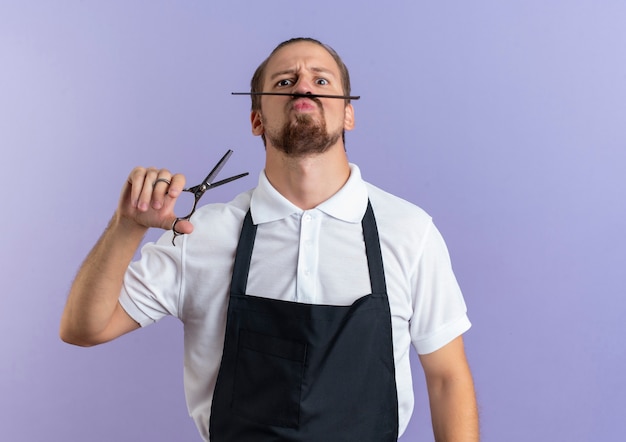 Drôle de jeune beau coiffeur immature portant uniforme tenant des ciseaux et tenant un peigne pour imiter sa fausse moustache isolée sur violet avec espace de copie