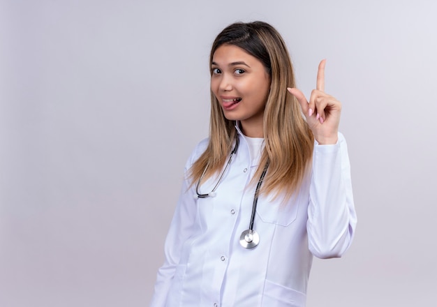 Drôle jeune belle femme médecin portant blouse blanche avec stéthoscope qui sort la langue pointant l'index vers le haut