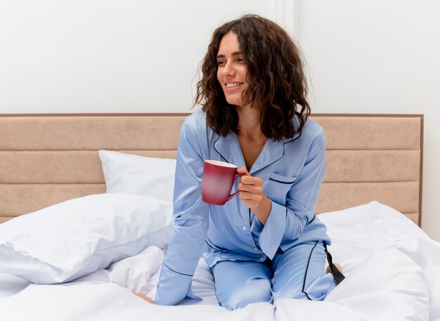 Drôle jeune belle femme en pyjama bleu assis sur le lit avec une tasse de café heureux et positif souriant gaiement à l'intérieur de la chambre sur fond clair