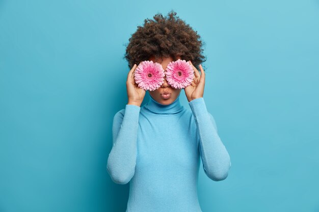 Drôle Jeune Femme Aux Cheveux Bouclés Couvre Les Yeux Avec La Marguerite Rose De Gerberas, Fait Le Bouquet Et Le Meilleur Cadeau Naturel Pour Un Ami, Porte Un Col Roulé Bleu. Carrière De Fleuriste. Belle Floraison, Parfum Agréable