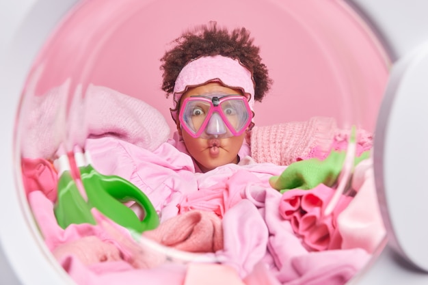 Photo gratuite une drôle de jeune femme aux cheveux bouclés fait la grimace des lèvres de poisson porte des poses de masque de plongée de l'intérieur de la machine à laver se prépare pour le processus de blanchiment entouré de tas de vêtements sales à laver