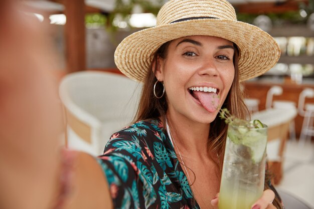 Drôle jeune femme à la mode montre la langue, fait un selfie avec un téléphone portable, tient un cocktail frais dans les mains, aime les loisirs et un bon repos d'été. Femme de touriste dans une cafétéria confortable