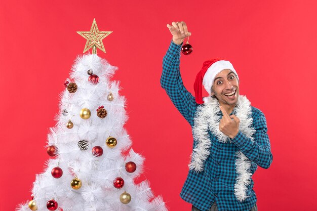 Drôle de jeune homme avec chapeau de père Noël dans une chemise dépouillé bleu et soulevant des accessoires de décoration montrant debout près de l'arbre de Noël sur le rouge