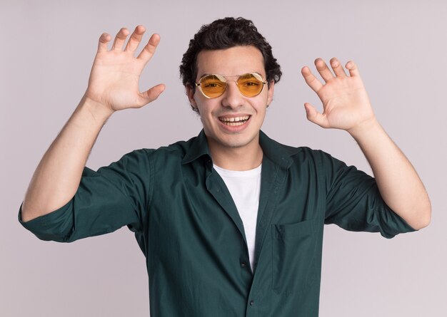 Drôle de jeune homme en chemise verte portant des lunettes à l'avant faisant le geste des griffes comme chat debout sur un mur blanc