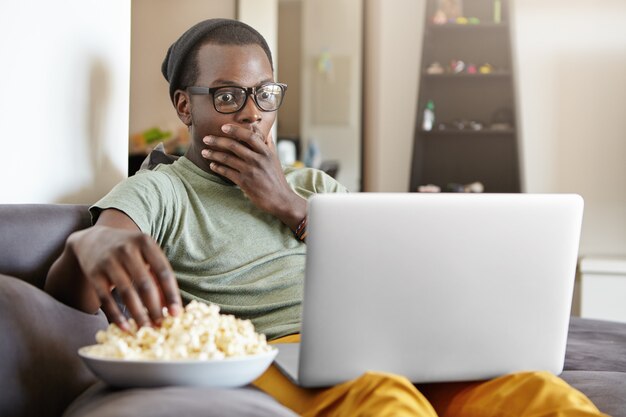 Drôle de jeune homme à la peau sombre assis sur un canapé gris dans le salon avec un ordinateur portable sur ses genoux, regardant l'écran avec un regard choqué ou effrayé, couvrant la bouche avec la main tout en regardant un film effrayant