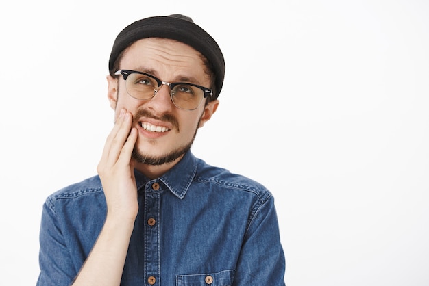 drôle mécontent mâle barbu sentiment d'inconfort grimaçant regardant et fronçant les sourcils de mal de dents douloureux tenant la main sur la joue et se plaignant de la douleur