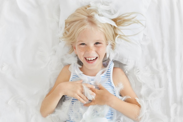 Drôle de petit enfant aux cheveux clairs, allongé sur des draps blancs, ressentant de la joie en attrapant des plumes, s'amusant avec ses amis.