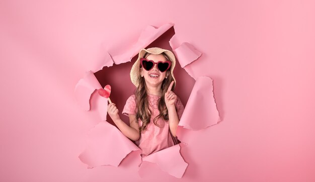 drôle de petite fille regarde par le trou dans un chapeau de plage et des lunettes en forme de coeur, tenant un coeur sur un bâton, sur un fond coloré, une place pour le texte, la prise de vue en studio