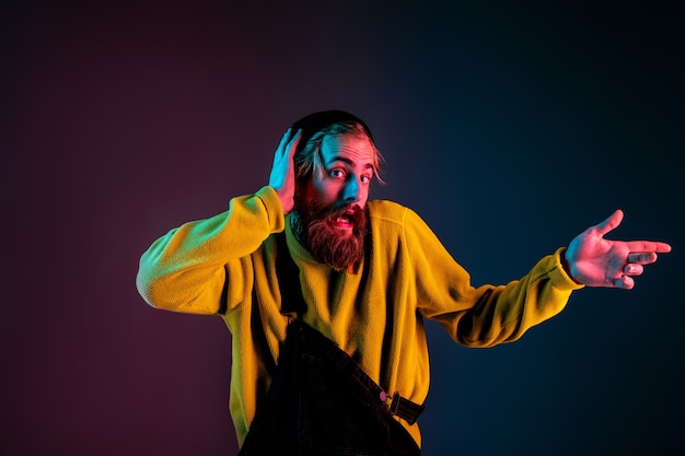 Drôle de pointage sur le côté. Portrait de l'homme caucasien sur fond de studio dégradé en néon. Beau modèle masculin avec un style hipster. Concept d'émotions humaines, expression faciale, ventes, publicité.