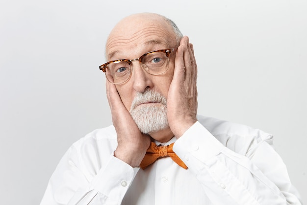 Photo gratuite drôle de vieil homme barbu dans des lunettes élégantes souffrant de terribles maux de dents, tenant les mains sur ses joues, bougeant ses yeux bleus. homme âgé effrayé exprimant le choc et l'étonnement