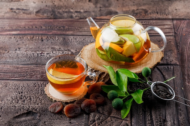 Eau infusée de fruits dans une théière avec des abricots séchés au thé, du bois, un récipient, des citrons verts vue grand angle sur une surface de carreaux de pierre