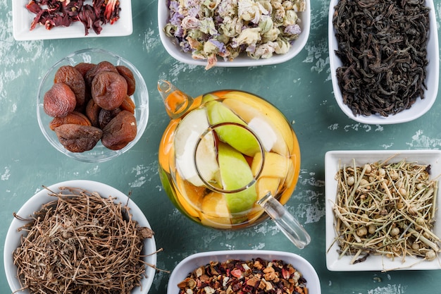 Eau infusée de fruits dans une théière avec des abricots secs, des herbes, des tiges de cerisier à plat sur une surface en plâtre