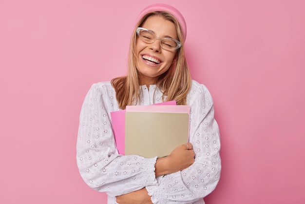 Une écolière ou un étudiant heureux, heureux de terminer la préparation de la session d'examen, tient des manuels, sourit largement, se sent joyeux, porte des lunettes transparentes et un chemisier blanc isolé sur rose