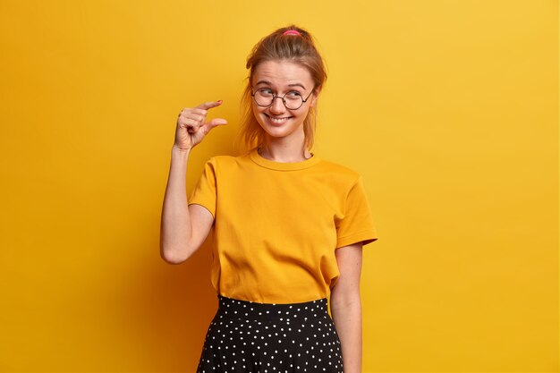 Une écolière intelligente et heureuse montre une petite quantité de quelque chose, mesure une toute petite chose, explique à quel point elle était intéressée pendant la leçon, porte des lunettes optiques