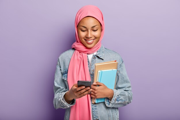 Une écolière musulmane ravie regarde des photos sur les réseaux sociaux, détient un cellulaire moderne, regarde du contenu vidéo amusant