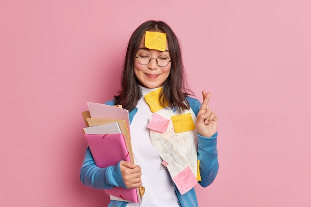 Une écolière positive croise les doigts croit en la bonne chance à l'examen porte des lunettes rondes collées avec des papiers et des notes autocollantes, des informations écrites à retenir font berceau. L'élève utilise des feuilles de triche.