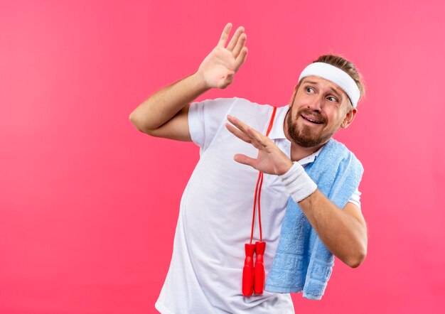 Effrayé jeune bel homme sportif portant un bandeau et des bracelets en levant les mains et en regardant de côté avec une corde à sauter et une serviette sur les épaules isolé sur l'espace rose