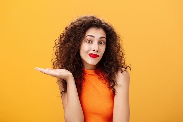 Eh bien désolé. Femme charmante et stupide inconsciente et désemparée avec une coiffure frisée, haussant les épaules et souriant avec un regard incertain levant la paume dans un geste qui sait, posant sans s'impliquer sur fond orange.