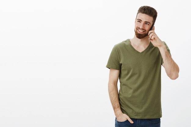 élégant et confiant beau entrepreneur masculin barbu parler sur smartphone souriant et riant appréciant la conversation en regardant le coin supérieur gauche sur le mur blanc