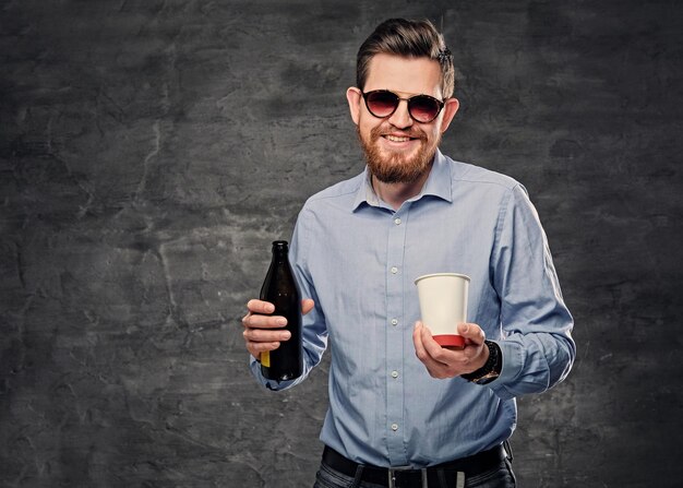 L'élégant hipster barbu tient une tasse de café en papier et de la bière artisanale en bouteille.