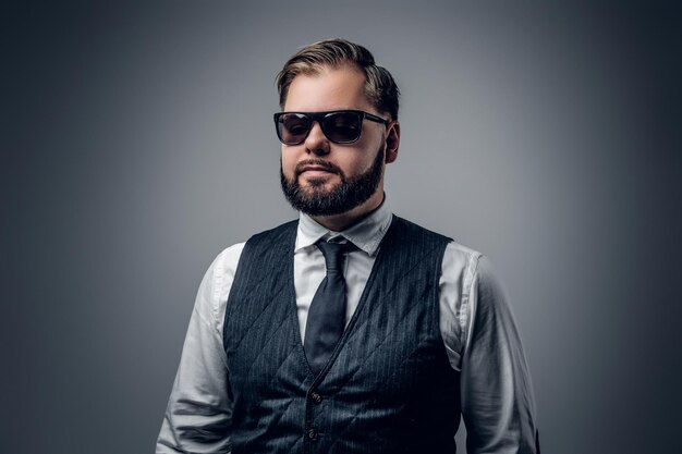 Un élégant homme d'affaires barbu portant des lunettes de soleil et un gilet sur fond gris.