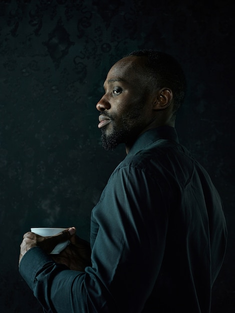 Photo gratuite Élégant jeune homme noir africain avec une tasse blanche de café posant sur fond sombre de studio.