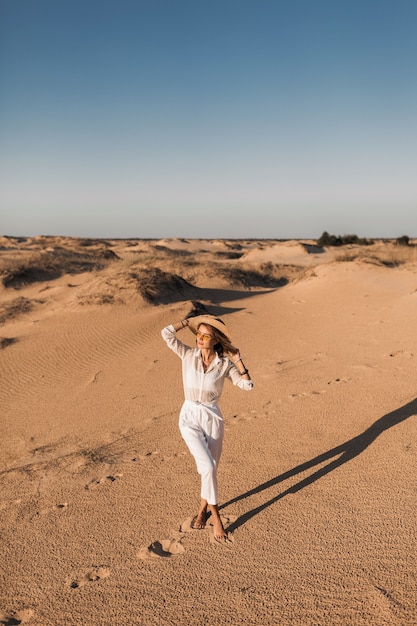 Photo gratuite Élégante belle femme marchant dans le sable du désert vêtue d'un pantalon blanc et d'un chemisier portant un chapeau de paille sur le coucher du soleil