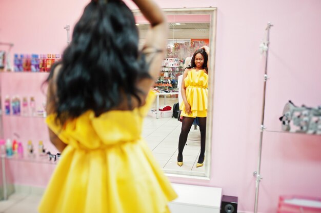 Élégante Femme Afro-américaine à La Robe Jaune Posée Dans Une Boutique Rose Contre Un Miroir