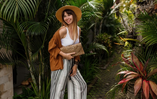 Photo gratuite Élégante fille élégante en haut blanc et chapeau de paille posant sur des feuilles de palmier à bali.