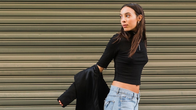 Photo gratuite Élégante jeune femme debout devant l'obturateur de fer regardant par-dessus l'épaule enlever la veste