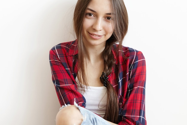 Photo gratuite Élégante à la mode jolie jeune femme vêtue de jeans déchiquetés et chemise à carreaux rouge sur haut blanc